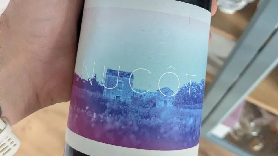 Bouteille de Nucôt du Clos Dauliac tenue en main, étiquette au dégradé bleu et violet représentant un mas en pierre